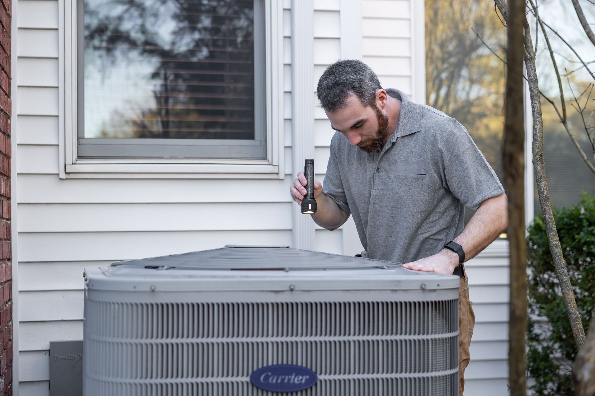 An HVAC technician inspecting an outdoor unit with a flashlight